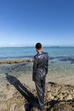 Model wearing a black wrap style sequin blazer with sunray stripes and matching wide leg trousers, standing on a rocky beach with clear blue water and sky.