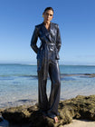 Model wearing a black wrap style sequin blazer with sunray stripes and matching wide leg trousers, standing on a rocky beach with clear blue water and sky.
