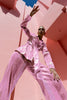 Model wearing a pink wrap-style sequin blazer with sunray stripes and matching wide-leg trousers, posing against a modern pink architectural background