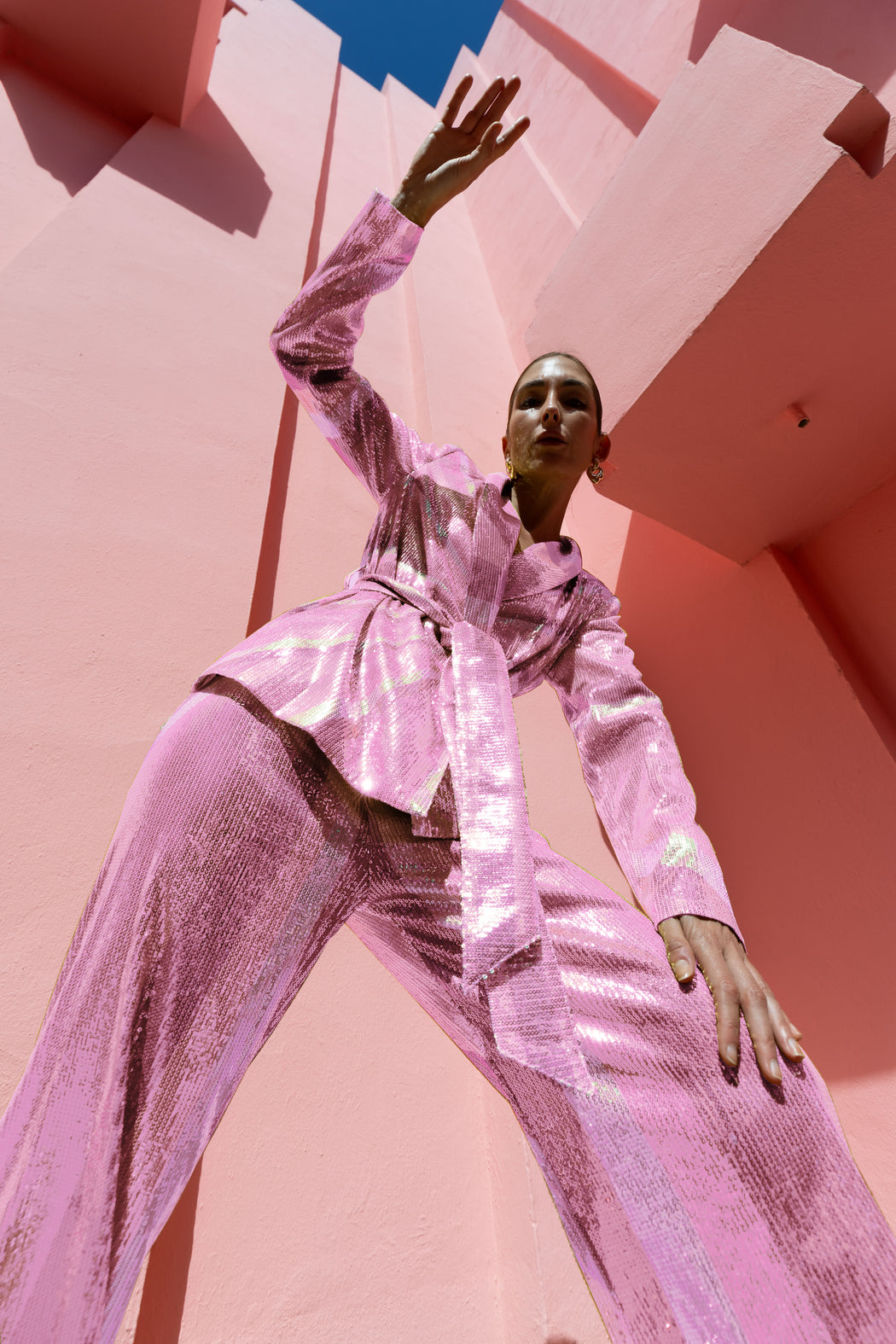Model wearing a pink wrap-style sequin blazer with sunray stripes and matching wide-leg trousers, posing against a modern pink architectural background