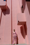 Model wearing a pink wrap-style sequin blazer with sunray stripes and matching wide-leg trousers posing against a geometric pink wall