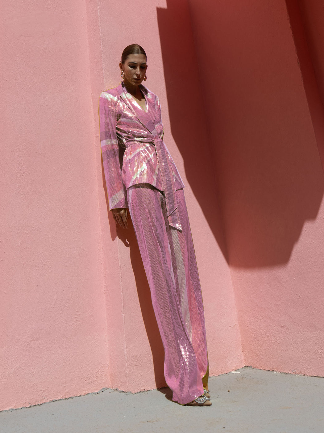 Model wearing a pink wrap-style sequin blazer with sunray stripes and matching wide-leg trousers lying against a pink wall