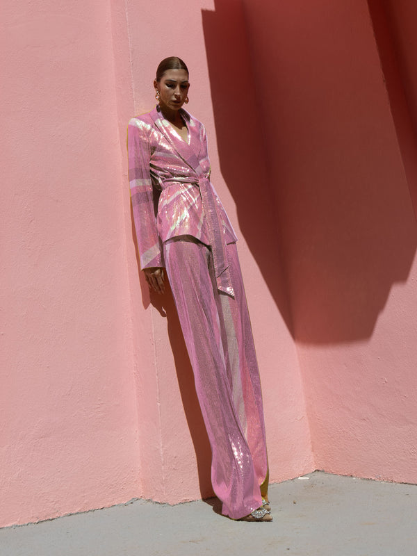 Model wearing a pink wrap-style sequin blazer with sunray stripes and matching wide-leg trousers lying against a pink wall