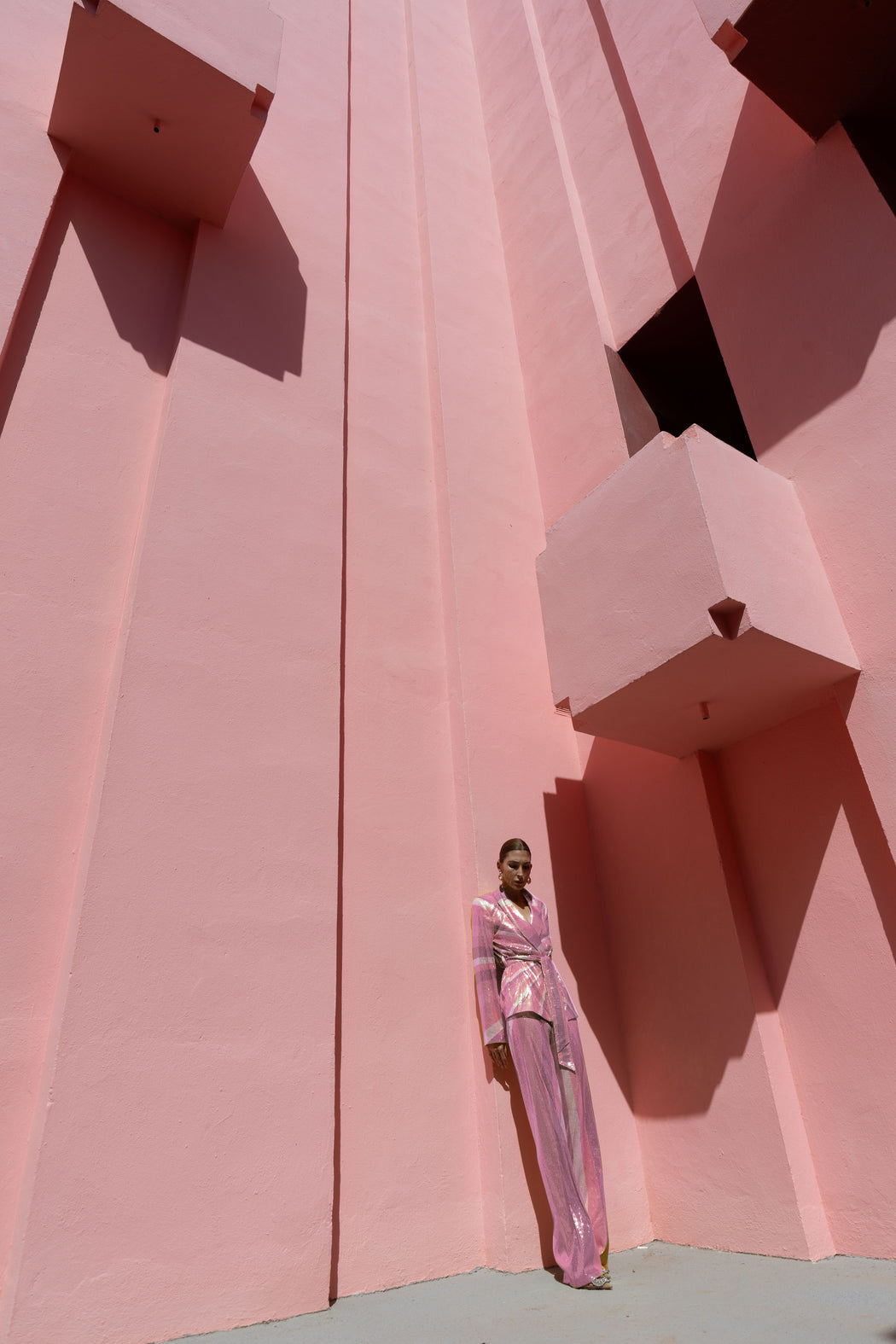 Model wearing a pink wrap-style sequin blazer with sunray stripes and matching wide-leg trousers lying against a geometric pink wall.