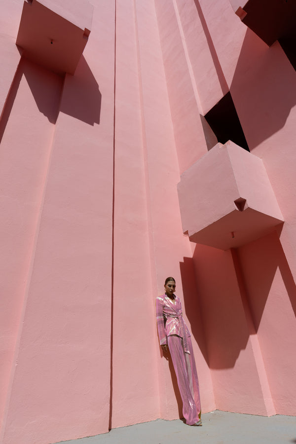 Model wearing a pink wrap-style sequin blazer with sunray stripes and matching wide-leg trousers lying against a geometric pink wall.