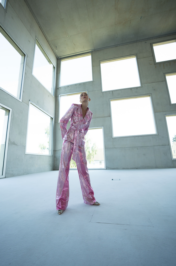 Model wearing a pink wrap-style sequin blazer with sunray stripes and matching wide-leg trousers standing in a modern indoor setting with large windows.