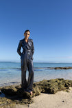 Model wearing a black wrap style sequin blazer with sunray stripes and matching wide leg trousers, standing on a rocky beach with clear blue water and sky.