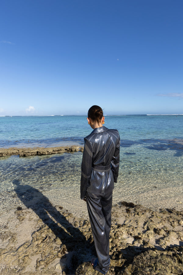 Model wearing a black wrap style sequin blazer with sunray stripes and matching wide leg trousers, standing on a rocky beach with clear blue water and sky.