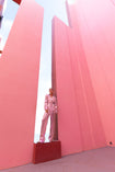 Model wearing a pink wrap-style sequin blazer with sunray stripes and matching wide-leg trousers standing between two tall pink structures with a clear blue sky.