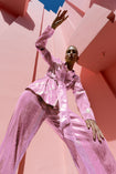 Model wearing a pink wrap-style sequin blazer with sunray stripes and matching wide-leg trousers, posing against a modern pink architectural background.