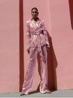 Model wearing a pink wrap-style sequin blazer with sunray stripes and matching wide-leg trousers, standing against a pink wall.