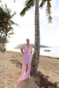 Model wearing a pink kaftan, elegantly pleated on certified recycled georgette fabric, with a gathered front waist, flowing sleeves, deep V-neckline, open back with button, and an included short slip dress, standing on a beach with palm trees and ocean in the background.