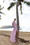Model wearing a pink kaftan, elegantly pleated on certified recycled georgette fabric, with a gathered front waist, flowing sleeves, deep V-neckline, open back with button, and an included short slip dress, standing on a beach with palm trees and mountains in the background.