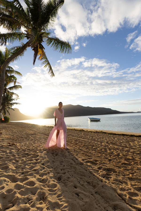 Model wearing a pink kaftan, elegantly pleated on certified recycled georgette fabric, with a gathered front waist, flowing sleeves, deep V-neckline, open back with button, and an included short slip dress, walking on a beach with palm trees and a boat in the background.
