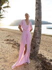 Model wearing a pink kaftan, elegantly pleated on certified recycled georgette fabric, with a gathered front waist, flowing sleeves, deep V-neckline, open back with button, and an included short slip dress, standing by a palm tree on a beach.