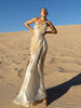 Model wearing an ivory metallic floor length dress with shimmer of gold pleated in a sunray pattern with circular shirring design and asymmetric neckline standing on sand dunes with a clear blue sky.