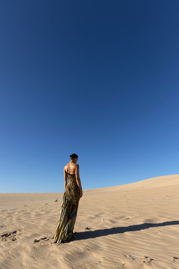 This is a photo of the back of the dress. Model standing on sand dunes wearing a floor-length, metallic gold dress with circular elastic shirring and a one-shoulder design. The dress features pleats in a Sunray pattern that contour the body and flatter the silhouette.