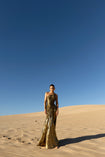 Model standing on sand dunes, wearing a floor-length, metallic gold dress with circular elastic shirring and a one-shoulder design. The dress features pleats in a Sunray pattern that contour the body and flatter the silhouette.