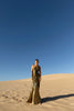 Model standing on sand dunes, wearing a floor-length, metallic gold dress with circular elastic shirring and a one-shoulder design. The dress features pleats in a Sunray pattern that contour the body and flatter the silhouette.
