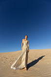 Model wearing an ivory metallic floor length dress with shimmer of gold pleated in a sunray pattern with circular shirring design and asymmetric neckline walking on sand dunes with a clear blue sky.
