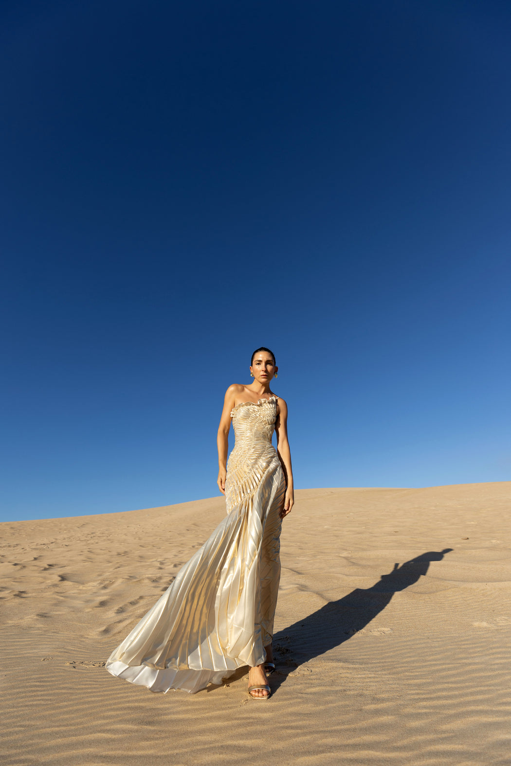 Model wearing an ivory metallic floor length dress with shimmer of gold pleated in a sunray pattern with circular shirring design and asymmetric neckline walking on sand dunes with a clear blue sky.