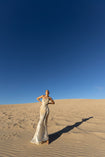 Model wearing an ivory metallic floor length dress with shimmer of gold pleated in a sunray pattern with circular shirring design and asymmetric neckline posing on sand dunes with a clear blue sky.