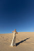 Model wearing an ivory metallic floor length dress with shimmer of gold pleated in a sunray pattern with circular shirring design and asymmetric neckline posing on sand dunes with a clear blue sky.
