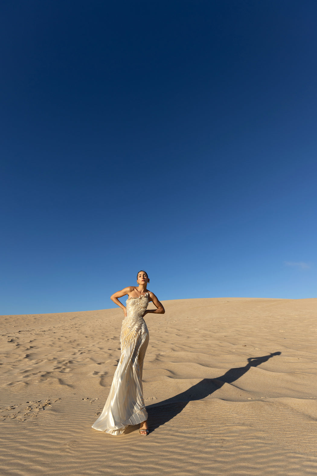 Model wearing an ivory metallic floor length dress with shimmer of gold pleated in a sunray pattern with circular shirring design and asymmetric neckline posing on sand dunes with a clear blue sky.