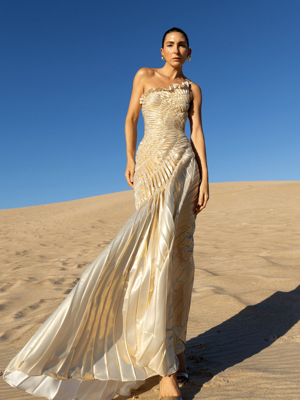 Model wearing an ivory metallic floor length dress with shimmer of gold pleated in a sunray pattern with circular shirring design and asymmetric neckline standing on sand dunes with a clear blue sky.