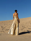 Model wearing an ivory metallic floor length dress with shimmer of gold pleated in a sunray pattern with circular shirring design and asymmetric neckline turned back while standing on sand dunes with a clear blue sky.