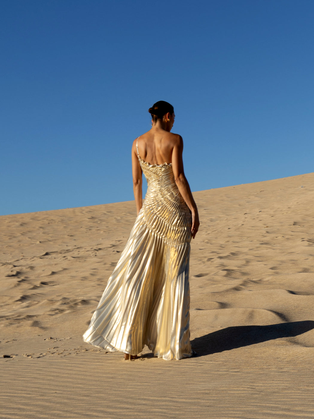 Model wearing an ivory metallic floor length dress with shimmer of gold pleated in a sunray pattern with circular shirring design and asymmetric neckline turned back while standing on sand dunes with a clear blue sky.