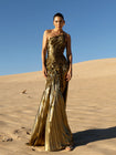 Model wearing a metallic gold sleeveless dress pleated in a sunray pattern with circular shirring design standing on sand dunes with a clear blue sky.