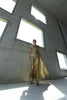 Model wearing a gold statement dress brought to life with all-over shimmering sequins. Intricate gathered tuck detailing in a circular motion creates a sculptural, flattering silhouette that catches the light beautifully from every angle, standing in a modern indoor setting with large windows.