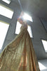 Model wearing a gold statement dress brought to life with all-over shimmering sequins. Intricate gathered tuck detailing in a circular motion creates a sculptural, flattering silhouette that catches the light beautifully from every angle, standing in a room with large windows.