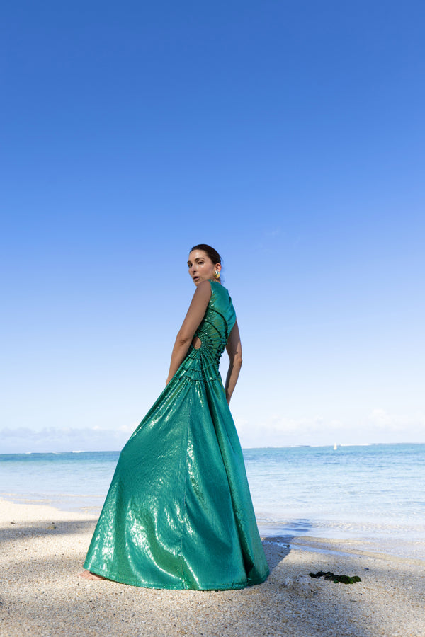 Model wearing a green statement dress adorned with all over shimmering sequins, featuring circular gathered tuck detailing that creates a sculptural silhouette, a sleeveless design with a round neckline, an artfully ruched bodice, and an adjustable side waist cut out. Standing on a beach with blue sky and ocean in the background.
