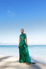 Model wearing a green statement dress adorned with all over shimmering sequins, featuring circular gathered tuck detailing that creates a sculptural silhouette, a sleeveless design with a round neckline, an artfully ruched bodice, and an adjustable side waist cut out. Standing on a beach with blue sky and ocean in the background.