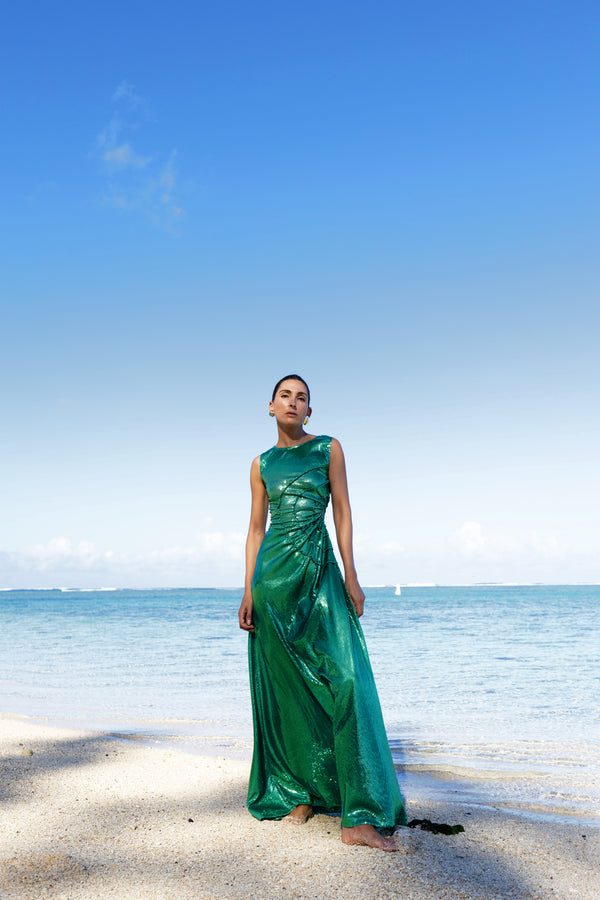 Model wearing a green statement dress adorned with all over shimmering sequins, featuring circular gathered tuck detailing that creates a sculptural silhouette, a sleeveless design with a round neckline, an artfully ruched bodice, and an adjustable side waist cut out. Standing on a beach with blue sky and ocean in the background.