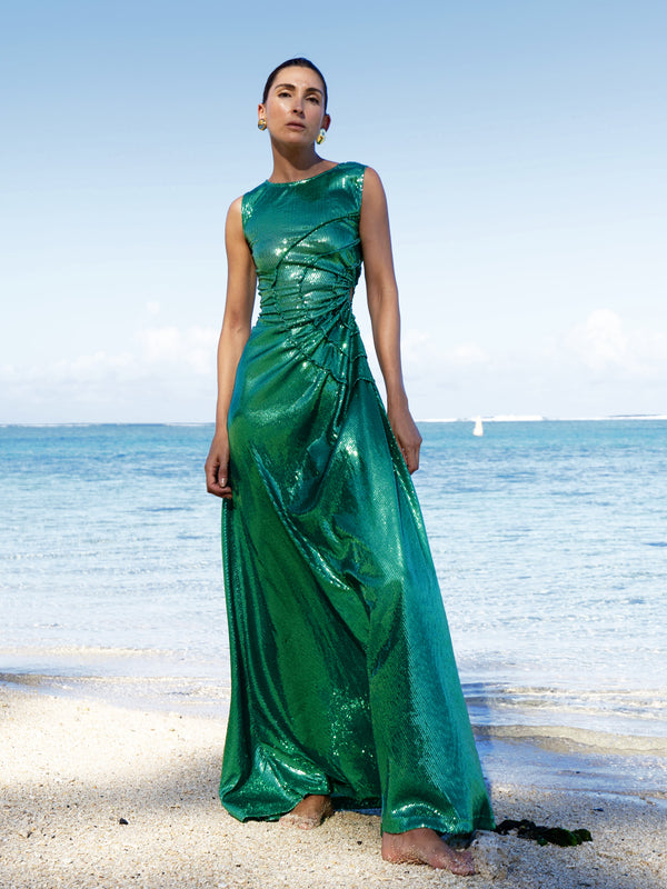 Model wearing a statement dress adorned with all over shimmering sequins, featuring circular gathered tuck detailing that creates a sculptural silhouette, a sleeveless design with a round neckline, an artfully ruched bodice, and an adjustable side waist cutout. Standing on a beach with ocean and sky in the background.