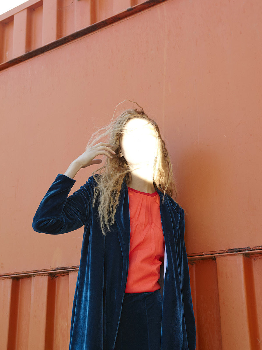 Model wearing a blue velvet coat over an orange shirt against a peach-coloured wall.