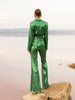 Model wearing a green sequin blazer with a wrap style front and exaggerated shawl lapel, paired with high rise green sequin trousers with a wide leg cut, creating a flattering, elongated, and statement making silhouette, standing on a rock with a beach background.