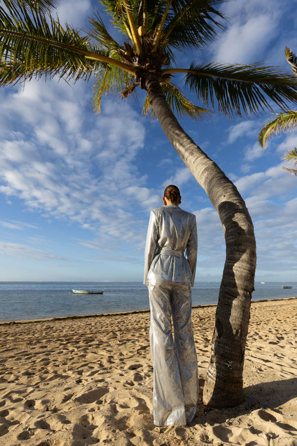 Model wearing a silver sequin blazer with a wrap style front and exaggerated shawl lapel, paired with high rise silver sequin trousers with a wide leg cut, creating a flattering, elongated, and statement making silhouette, standing on a beach with palm trees and ocean view.