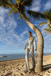 Model wearing a silver sequin blazer with a wrap style front and exaggerated shawl lapel, paired with high rise silver sequin trousers with a wide leg cut, creating a flattering, elongated, and statement making silhouette, standing on a beach with palm trees and ocean in the background.