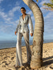Model wearing a silver sequin blazer with a wrap style front and exaggerated shawl lapel, paired with high rise silver sequin trousers with a wide leg cut, creating a flattering, elongated, and statement making silhouette, standing on a beach with palm trees and ocean in the background.