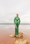 Model wearing a green sequin blazer with a wrap style front and exaggerated shawl lapel, paired with high rise green sequin trousers with a wide leg cut, creating a flattering, elongated, and statement making silhouette, standing on a rock by a body of water with a cloudy sky.