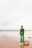 Model wearing a green sequin blazer with a wrap style front and exaggerated shawl lapel, paired with high rise green sequin trousers with a wide leg cut, creating a flattering, elongated, and statement making silhouette, standing on a rock by a body of water with a pastel gradient sky.