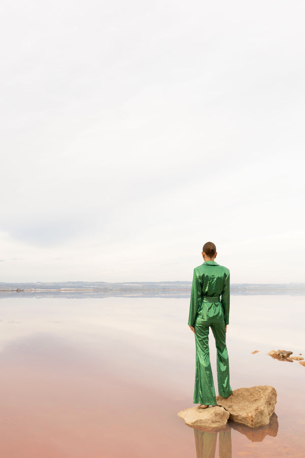 Model wearing a green sequin blazer with a wrap style front and exaggerated shawl lapel, paired with high rise green sequin trousers with a wide leg cut, creating a flattering, elongated, and statement making silhouette, standing on a rock by a body of water with a pastel gradient sky.