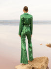 Model wearing a green sequin blazer with a wrap style front and exaggerated shawl lapel, paired with high rise green sequin trousers with a wide leg cut, creating a flattering, elongated, and statement making silhouette, standing on a rock with a beach background.