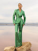 Model wearing a green sequin blazer with a wrap style front and exaggerated shawl lapel, paired with high rise green sequin trousers with a wide leg cut, creating a flattering, elongated, and statement making silhouette, standing on a rock with a beach background.