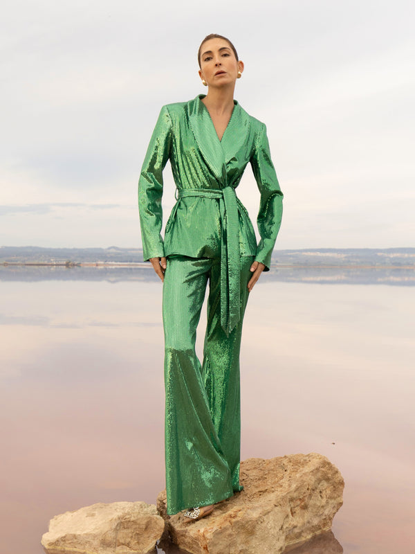 Model wearing a green sequin blazer with a wrap style front and exaggerated shawl lapel, paired with high rise green sequin trousers with a wide leg cut, creating a flattering, elongated, and statement making silhouette, standing on a rock with a beach background.