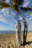 Model wearing a silver sequin blazer with a wrap style front and exaggerated shawl lapel, paired with high rise silver sequin trousers with a wide leg cut, creating a flattering, elongated, and statement making silhouette, standing on a beach with palm trees and ocean in the background.
